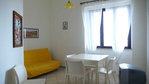 A small, bright room with a yellow sofa, a white table with chairs, a ceiling light, and a window with sheer curtains. - ARBACANI HOLIDAYS (Gallipoli)