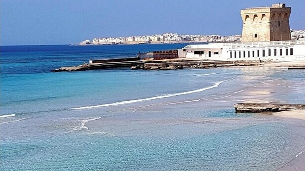 A coastal scene with clear blue waters, a sandy beach, and a historic building with a tower. - ARBACANI HOLIDAYS (Gallipoli)