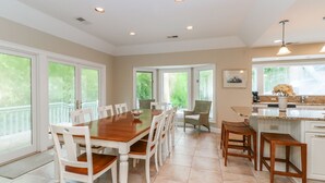 A spacious dining area with a large wooden table and white chairs, a kitchen with a marble countertop, and large windows offering a view of greenery. - DOG FRIENDLY Four Bedroom 3rd Row Ocean Sea Pines Home with a Private Pool (Hilton Head Island)