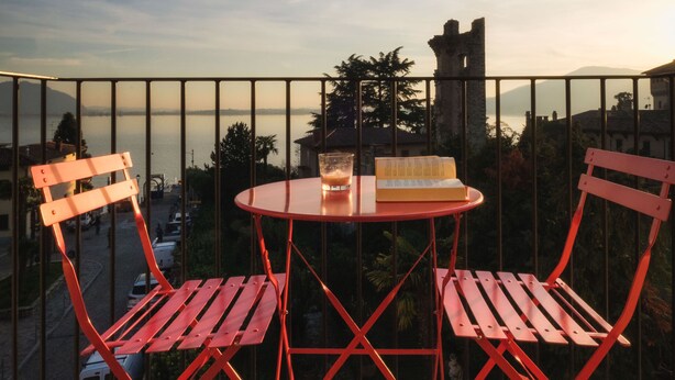 Ein Balkon mit einem roten Metalltisch und Stühlen, einem Getränk und einem Buch. Im Hintergrund ist ein Gewässer und ein Gebäude mit einem Turm zu sehen.