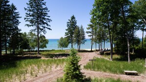Un terrain de camping avec un sentier de terre menant à un banc, entouré de grands arbres et avec une étendue d'eau en arrière-plan.