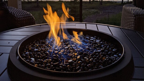 Un foyer extérieur circulaire, avec des flammes, entouré d’une rambarde métallique, et offrant une vue sur une étendue d’eau et des arbres.