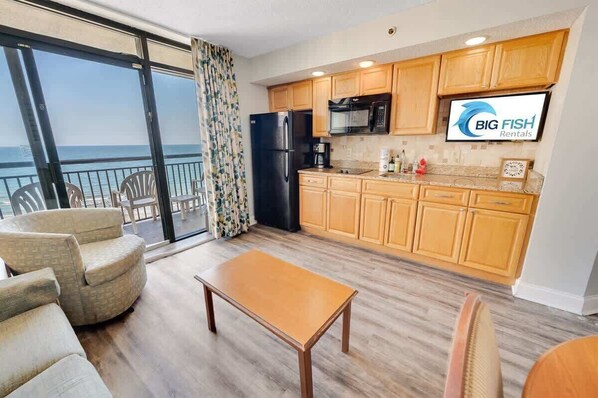 A modern kitchen with wooden cabinets, a black refrigerator, and a microwave. There is a dining area with a wooden table and chairs. The room has large windows with a view of the ocean. - New Beachfront, Condo Simply Amazing Pools & Views (North Myrtle Beach)