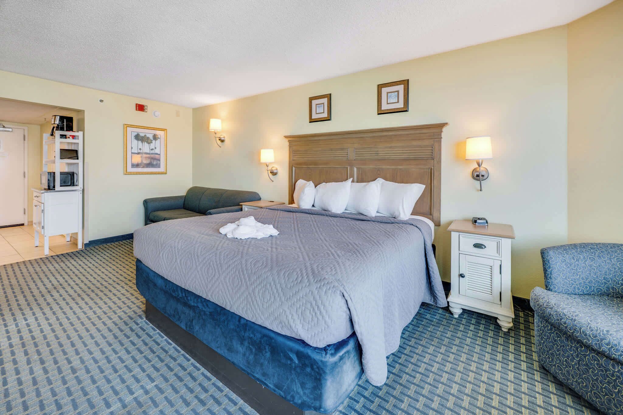 A hotel room with a bed, bedside tables, a chair, a desk with a microwave, and a door leading to another room.