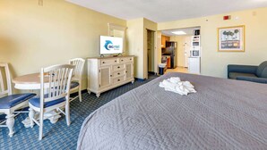 A hotel room with a bed, a desk with a chair, a dresser, a television, and a kitchenette in the background.
