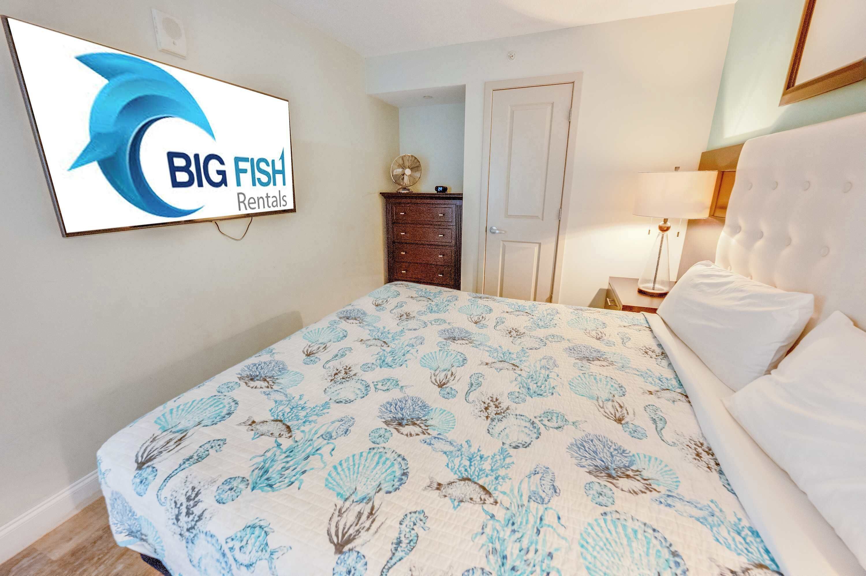 A hotel room with a bed, a dresser, a lamp, and a wall-mounted TV displaying 'BIG FISH Rentals'.