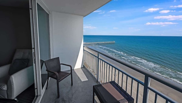 Ein Balkon mit Blick auf das Meer und einen Steg. Auf dem Balkon befinden sich ein Stuhl und ein kleiner Tisch.