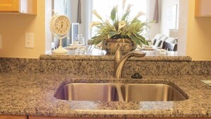 A kitchen with granite countertops, a stainless steel sink, a clock, and a potted plant. - Windsor_palms_303cp-n (Kissimmee)