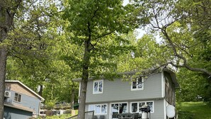 Une maison dotée d’une terrasse, entourée d’arbres et bordée d’une pelouse. On y trouve des chaises Adirondack et un foyer en métal.