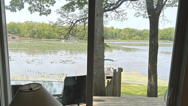 Vue depuis l’intérieur d’une pièce sur une terrasse en bois donnant sur un lac, avec des arbres et un bateau amarré.