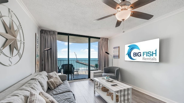 A modern living room with a sofa, a coffee table, a ceiling fan, and a large window with a view of the ocean. - WOW Perfect Honeymoon Getaway with Great Views (Myrtle Beach)