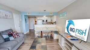 A modern living room with a grey sofa, a coffee table, and a dining area with white chairs. A kitchen with white cabinets and a microwave is visible in the background. A sign for 'Big Fish Rentals' is on the wall. - Sand Dunes Resort, Ocean View Getaway, 5th  Floor! (Myrtle Beach)