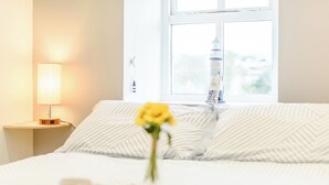A neatly made bed with striped pillows, a bedside lamp, a small table with a candle and a flower, and a view of a lighthouse through a window.