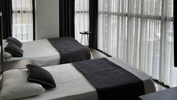 Una habitación de hotel con dos camas, una mesita pequeña y ventanas amplias con cortinas traslúcidas.