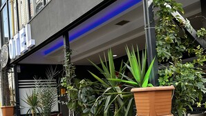 Entrada de hotel con un letrero, plantas en macetas y mesas al aire libre.