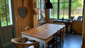 Een eetgedeelte met een houten tafel en rieten stoelen, grote ramen met uitzicht op groen en een hangende hanglamp.