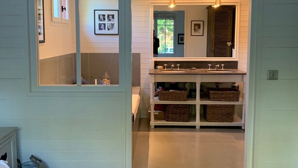 Een slaapkamer met een bed, een dressoir, een spiegel en een deur naar een badkamer met een wastafel en planken.