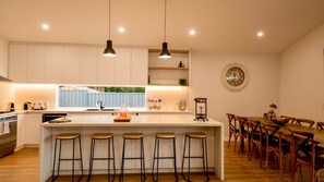 Cozinha moderna com ilha central, banquetas, área de refeições com cadeiras de madeira e um relógio na parede.