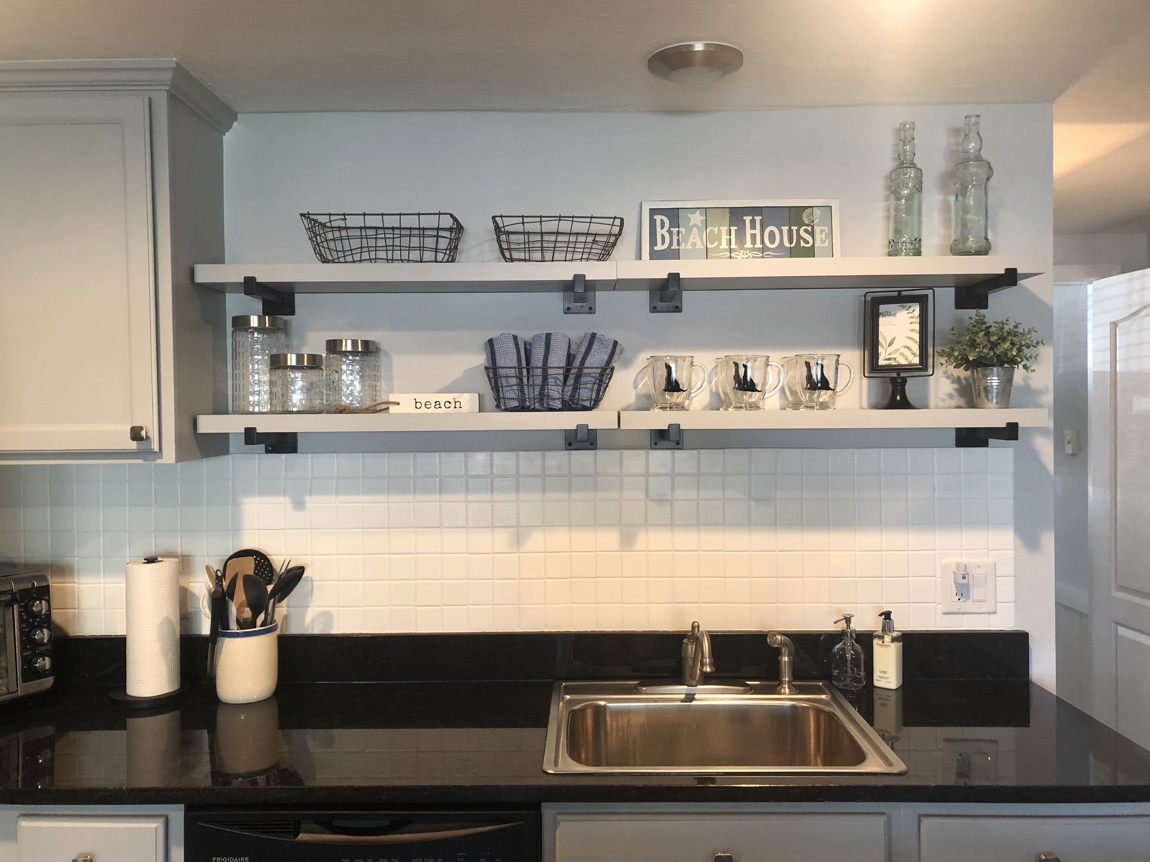 A kitchen with white cabinets, a black countertop, a stainless steel sink, and decorative items on shelves.