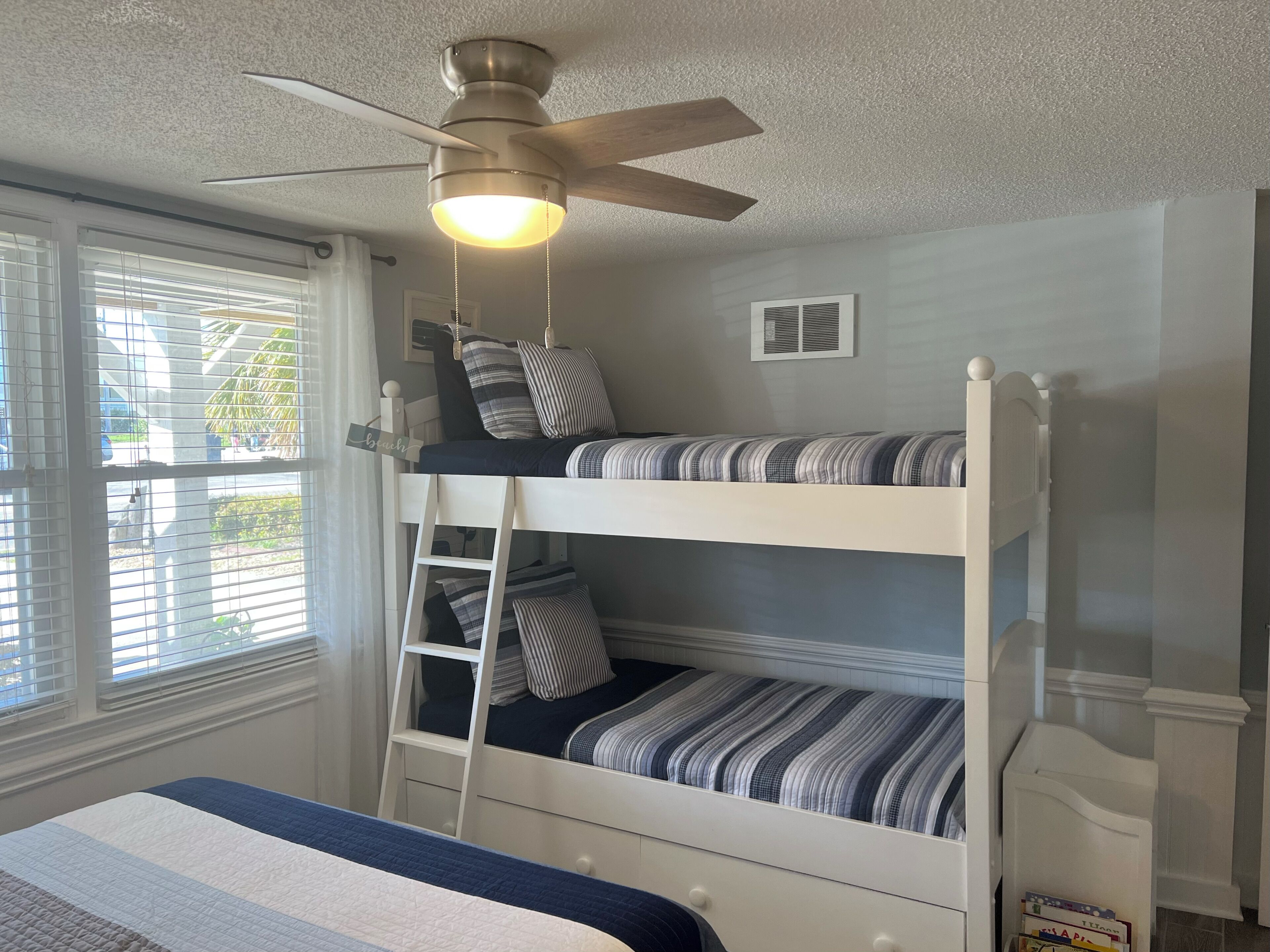 Une chambre avec un lit superposé, un ventilateur de plafond, une fenêtre avec des stores et une petite grille d’aération sur le mur.