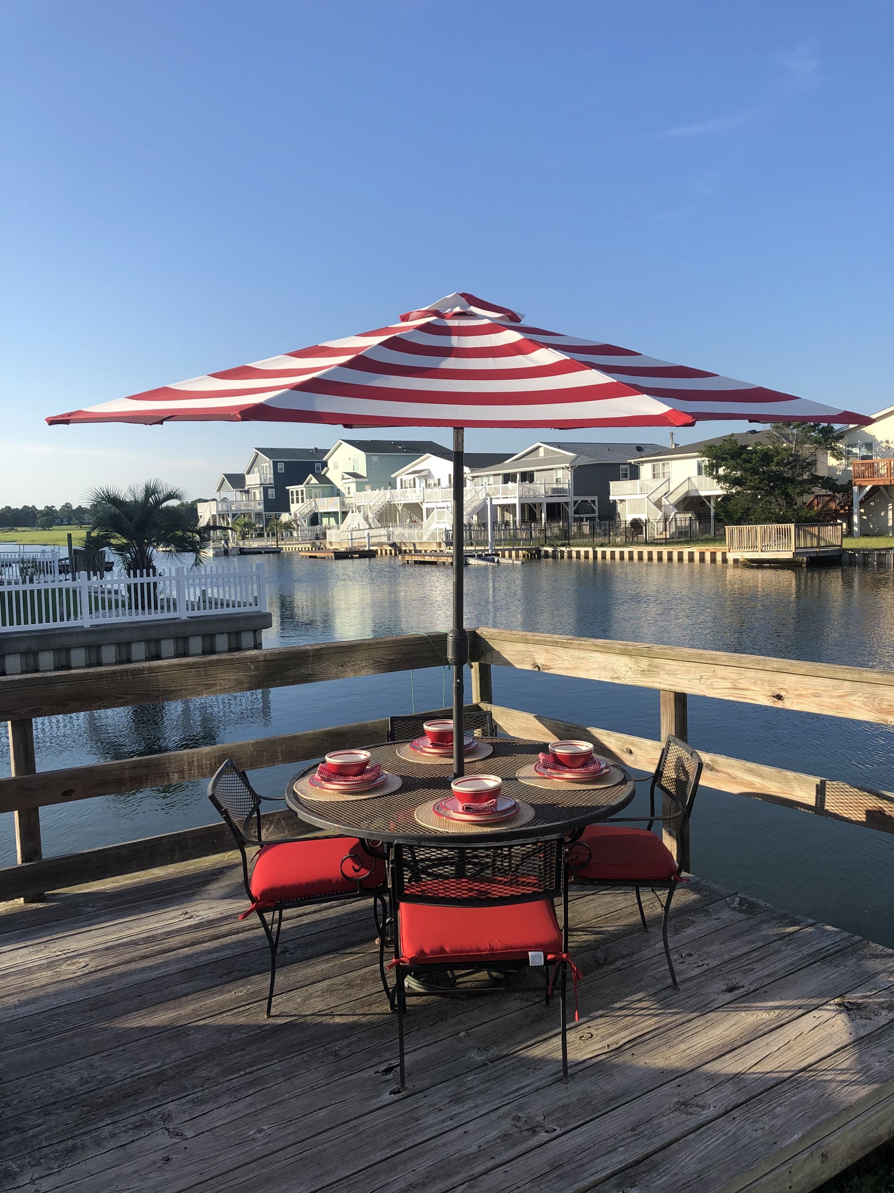 A wooden deck with a table and chairs under a striped umbrella, overlooking a body of water with houses in the background.