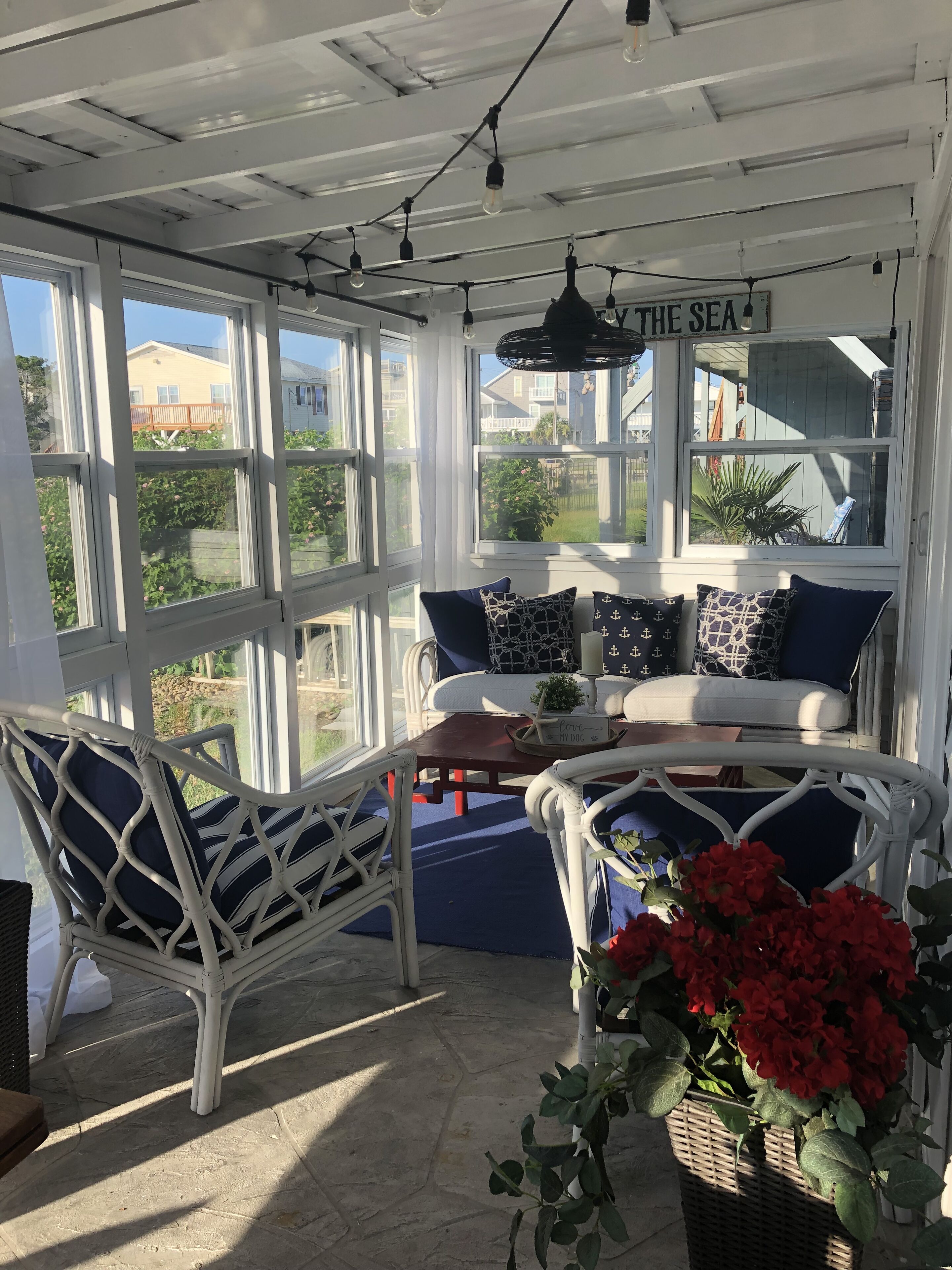 A sunlit room with a view of the sea, featuring a white wicker sofa with blue cushions, a wicker chair, and a small table with a plant.