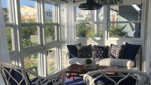 Et solbadet værelse med havudsigt, med en hvid fletsofa med blå puder, en fletstol og et lille bord med en plante.