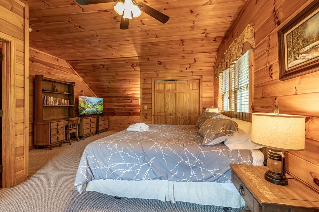 Une chambre confortable dans un chalet en rondins, avec un grand lit, une télévision, une bibliothèque et un ventilateur de plafond en bois.