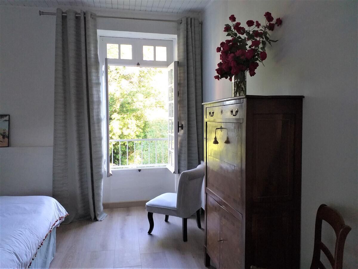 A bedroom with a bed, a chair, a wooden wardrobe, a window with curtains, and a vase with flowers on the wall.