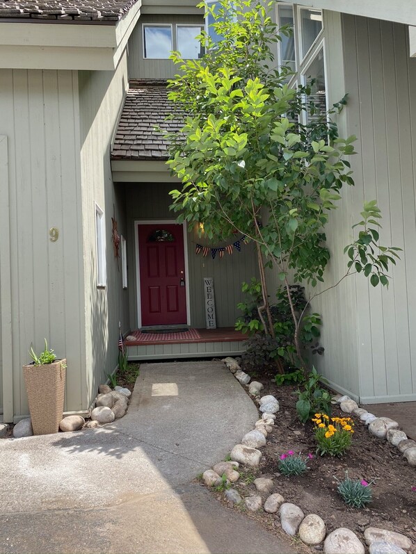 A house with a red door, a welcome sign, a small garden with rocks and flowers, and a potted plant. - Free Shuttle to Grand Targhee! Stay at the Powder Valley Condo, welcome basket! (Driggs)