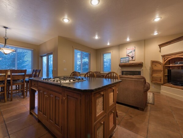 Una cocina amplia con una isla central, armarios de madera y una estufa empotrada. Varias ventanas ofrecen luz natural y un rincón acogedor con chimenea.