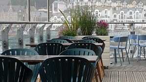 A terrace with wooden flooring and metal railings, featuring a table with chairs and potted plants.