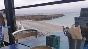 A table with a cup of coffee and a spoon, a metal bucket with utensils, and a view of a beach and sea through a window.