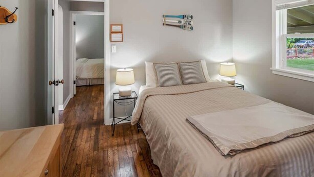 A bedroom with a wooden floor, a bed with a striped pillow, two bedside lamps, and a window with a view of a garden. - Lake Street 425 ~ Fantastic 2bed/2bath Home Right (Sandpoint)