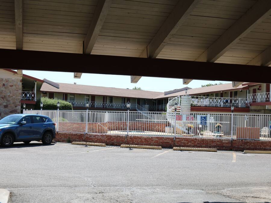 A parking area with a blue car, a covered walkway, and a building with a balcony and a fenced area.