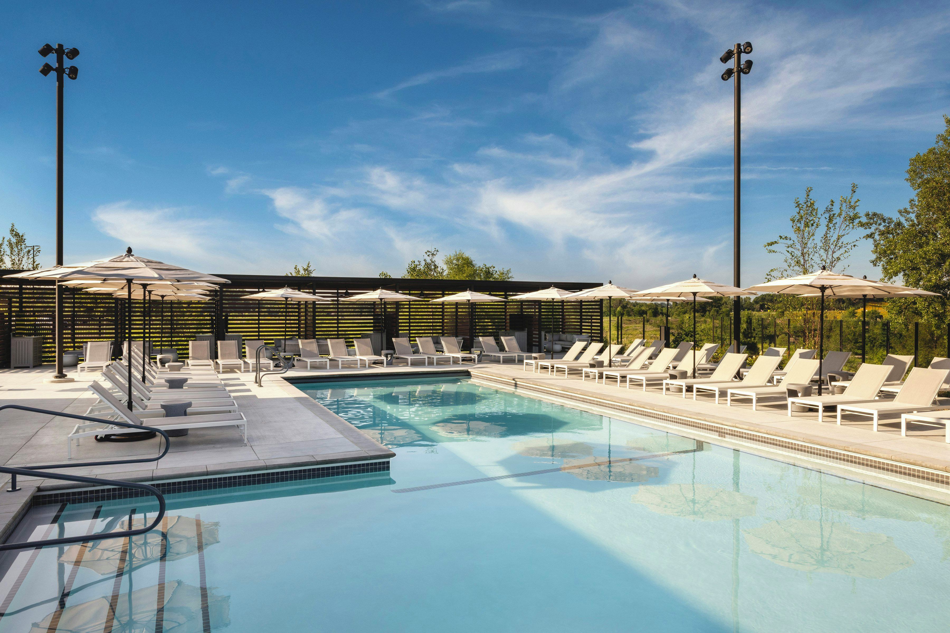 Ein Swimmingpool mit Liegestühlen und Sonnenschirmen, umgeben von Bäumen und blauem Himmel.