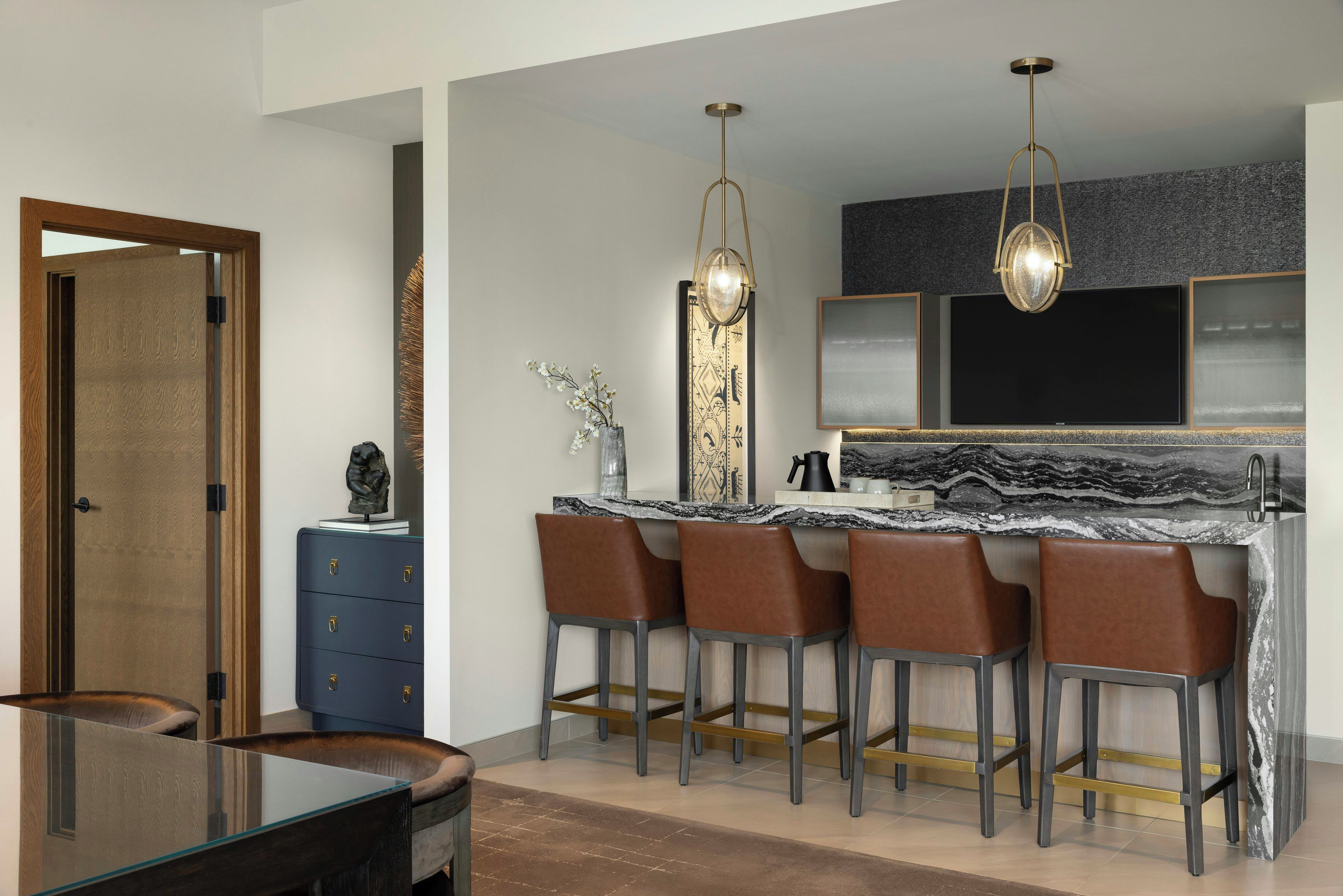 A modern kitchen with a marble countertop, bar stools, pendant lighting, and a large mirror above the sink.