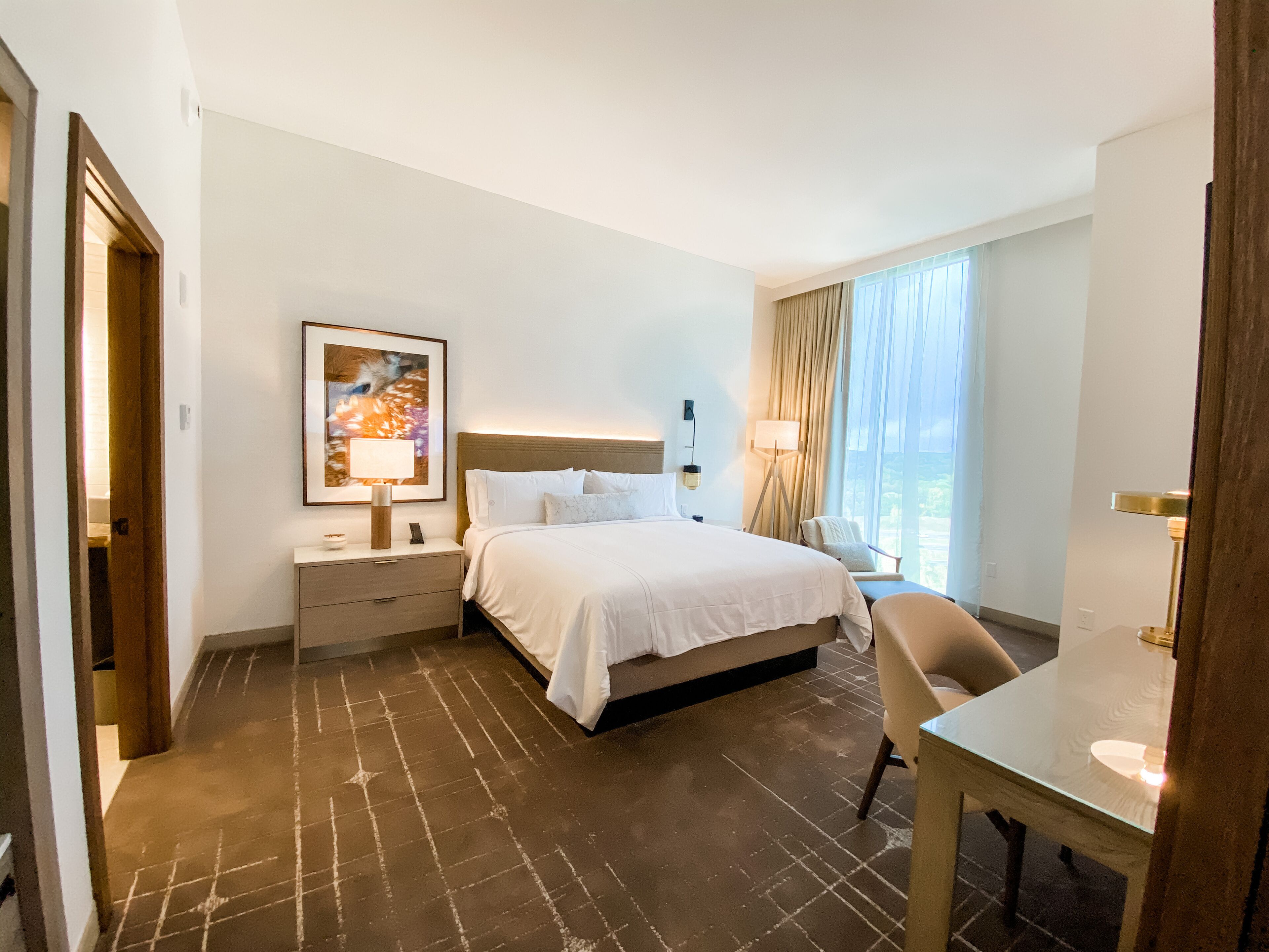 A hotel room with a bed, bedside table, lamp, chair, and a large window with curtains.