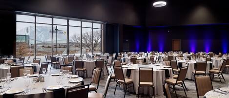 Un amplio salón de banquetes con mesas redondas dispuestas para comer, grandes ventanales y vistas al exterior.