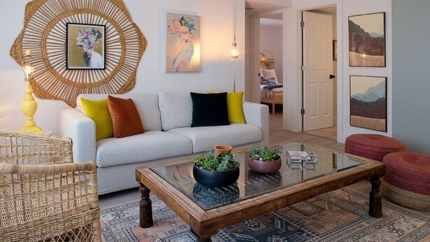 A living room with a white sofa, colorful cushions, a glass coffee table with plants, a woven wall hanging, and a patterned rug. - Stylish & Eclectic Modern 2/2 Apt - WeArePeaceful (South Pasadena)
