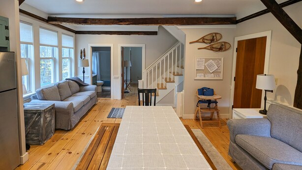 A cozy living room with a wooden floor, a sofa, a dining table, and a staircase leading to another level. - 1810 Brook Side Mill/House / EV Level 2 Charging on Site (Shelburne Falls)