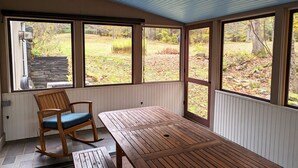 A covered patio with a wooden table and a rocking chair, large windows offering a view of a garden. - 1810 Brook Side Mill/House / EV Level 2 Charging on Site (Shelburne Falls)