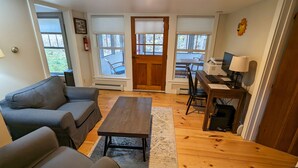 A living room with a sofa, a coffee table, and a desk with a computer. There are windows with blinds, a door, and a fire extinguisher on the wall. - 1810 Brook Side Mill/House / EV Level 2 Charging on Site (Shelburne Falls)