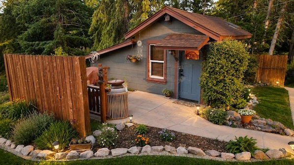 A small house with a wooden deck, a front door, and a garden with plants and a stone pathway.