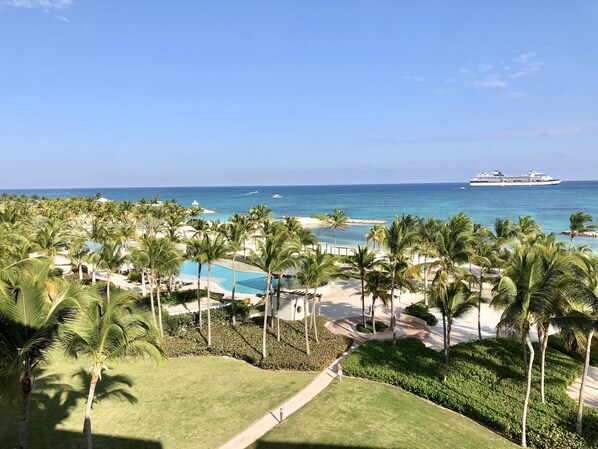 A resort with a swimming pool, palm trees, and a view of the ocean and a cruise ship.