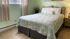 A bedroom with a bed, a nightstand, a lamp, a painting of a beach scene, and a window with curtains. - 1BR Condo Beach Block Location – Under 500 feet to the Beach & Boardwalk (Ocean City)