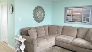 A living room with a beige sectional sofa, a small white side table, a decorative mirror, and a ceiling fan. - 1BR Condo Beach Block Location – Under 500 feet to the Beach & Boardwalk (Ocean City)