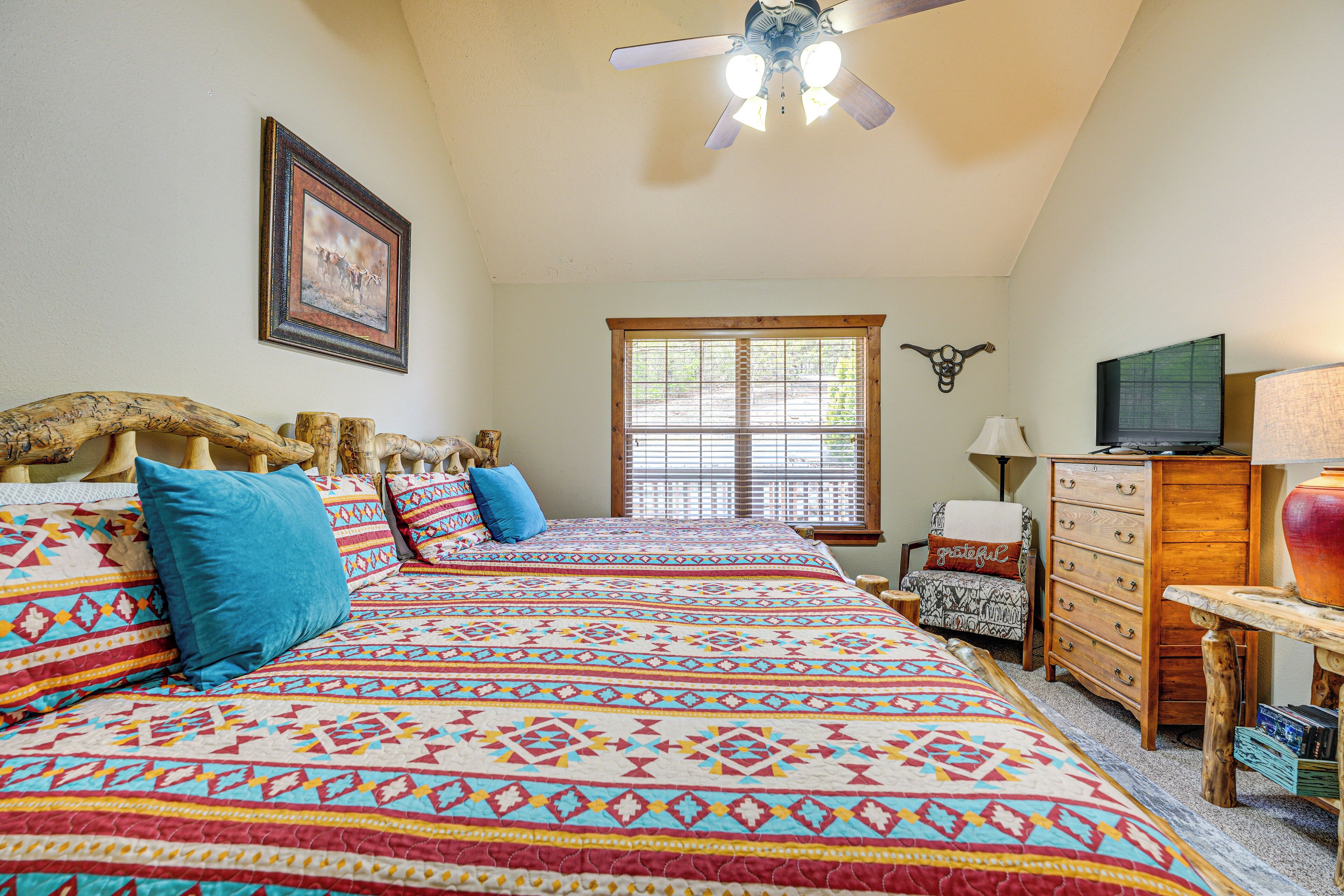 A bedroom with a large bed, a ceiling fan, a window with blinds, a dresser, and a television.