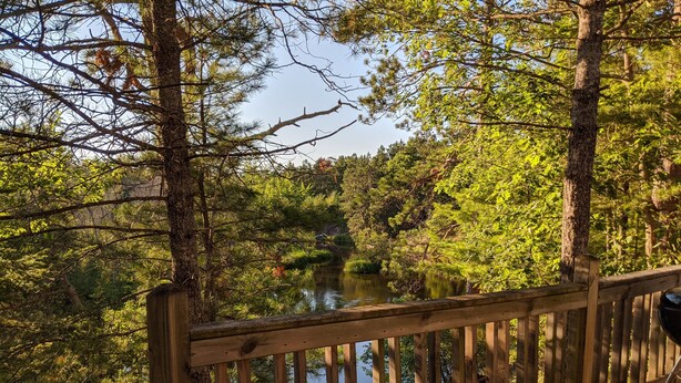 A wooden deck overlooking a serene lake surrounded by dense forest. - Treehouse on the river. (Au Train)