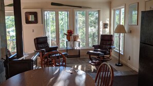 A spacious living room with large windows, a ceiling fan, and a dining area with a table and chairs. - Treehouse on the river. (Au Train)